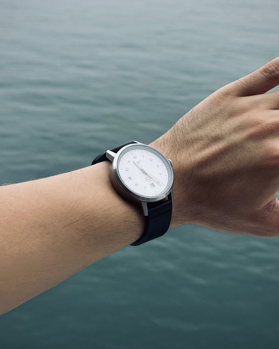 A minimalist analog wristwatch with a silver-toned case, white dial, and black strap worn on a person's wrist, with a blurred body of water in the background | White dial automatic watches V-Apostle with Black Horween Leather Straps | Vstelle Watch
